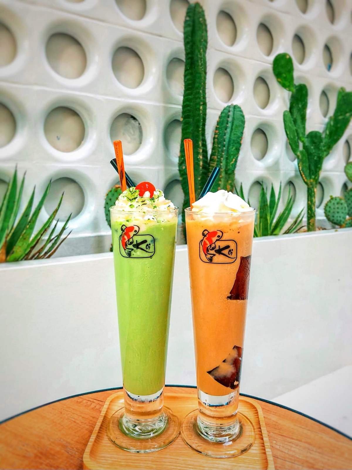 Hai ly sinh tố m&agrave;u xanh v&agrave; cam đặt tr&ecirc;n b&agrave;n gỗ, nền tường c&oacute; c&acirc;y xương rồng trang tr&iacute;.