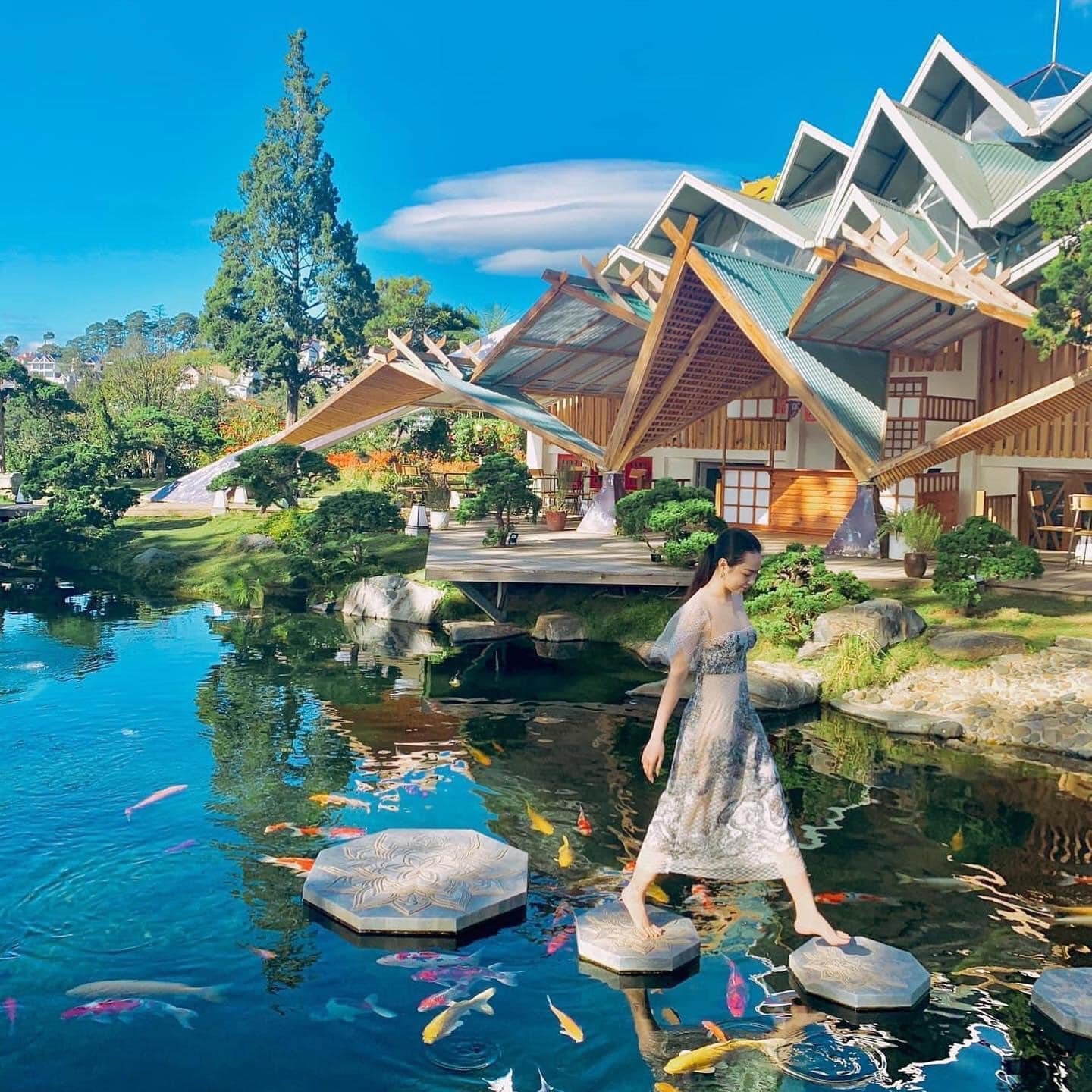 Người phụ nữ đi tr&ecirc;n c&aacute;c phiến đ&aacute; nổi giữa hồ c&aacute; Koi, b&ecirc;n cạnh l&agrave; ng&ocirc;i nh&agrave; c&oacute; kiến tr&uacute;c độc đ&aacute;o v&agrave; c&acirc;y xanh.