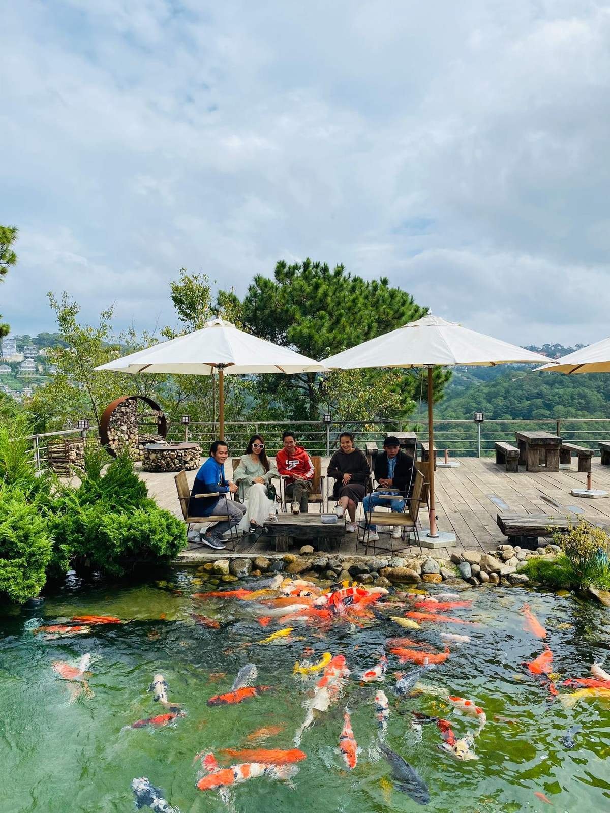 Nh&oacute;m người ngồi dưới d&ugrave; trắng b&ecirc;n hồ c&aacute; koi với cảnh quan c&acirc;y xanh v&agrave; n&uacute;i non ph&iacute;a xa.