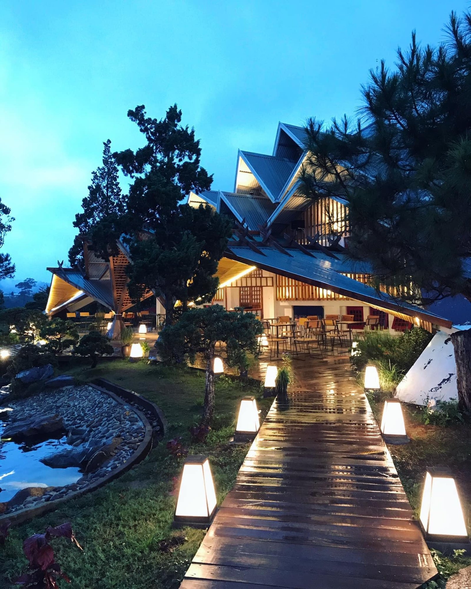 Lối đi gỗ dẫn đến t&ograve;a nh&agrave; c&oacute; m&aacute;i độc đ&aacute;o, xung quanh l&agrave; c&acirc;y xanh v&agrave; đ&egrave;n chiếu s&aacute;ng v&agrave;o l&uacute;c chạng vạng.