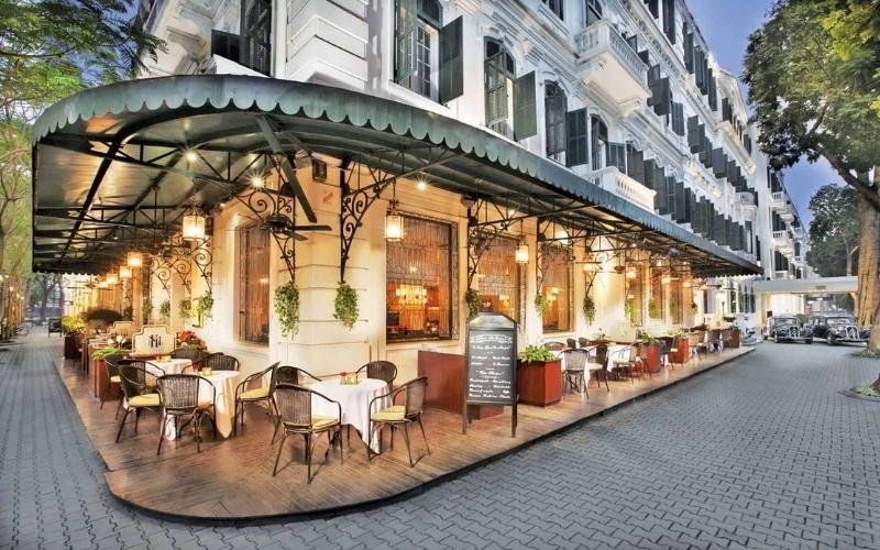 Quán cà phê ngoài trời với bàn ghế sắp xếp gọn gàng, nằm bên cạnh tòa nhà lớn phong cách cổ điển.