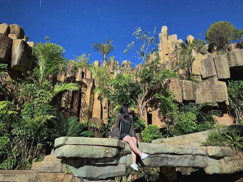 Người ngồi tr&ecirc;n tảng đ&aacute; lớn, ph&iacute;a sau l&agrave; cảnh n&uacute;i đ&aacute; v&agrave; c&acirc;y cối xanh tươi dưới bầu trời xanh.