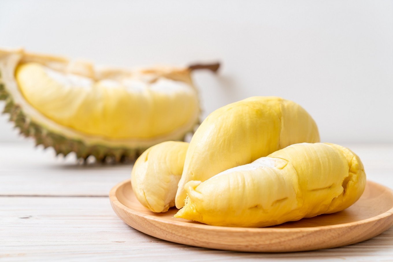Miếng sầu riêng vàng trên đĩa gỗ, phía sau là vỏ sầu riêng đã bổ, nền trắng.