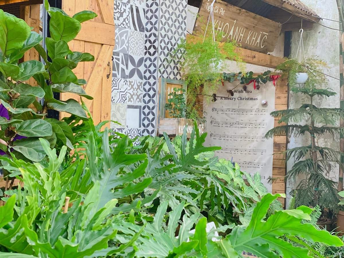 Qu&aacute;n c&agrave; ph&ecirc; ngo&agrave;i trời với c&acirc;y xanh tươi tốt, tường gạch hoa văn v&agrave; bảng nhạc Gi&aacute;ng Sinh trang tr&iacute;.