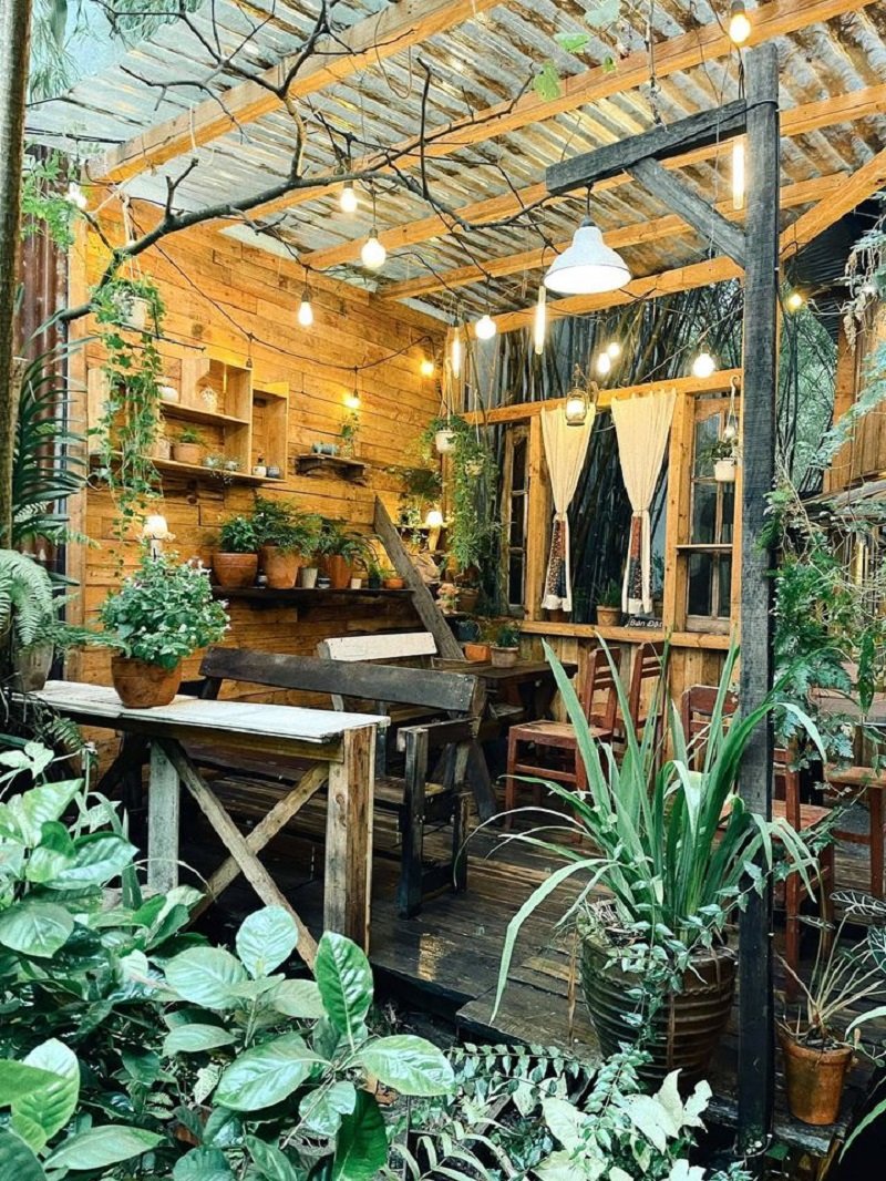Qu&aacute;n c&agrave; ph&ecirc; ngo&agrave;i trời với nội thất gỗ, c&acirc;y xanh v&agrave; đ&egrave;n treo trang tr&iacute;, tạo kh&ocirc;ng gian ấm c&uacute;ng v&agrave; thư gi&atilde;n.
