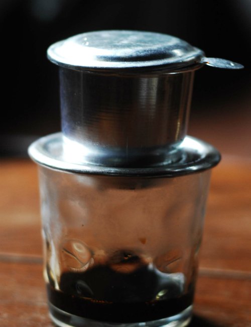 lycafe-9385-1435799050 Phin cà phê kim loại đang pha cà phê đen trên ly thủy tinh, đặt trên bề mặt gỗ.