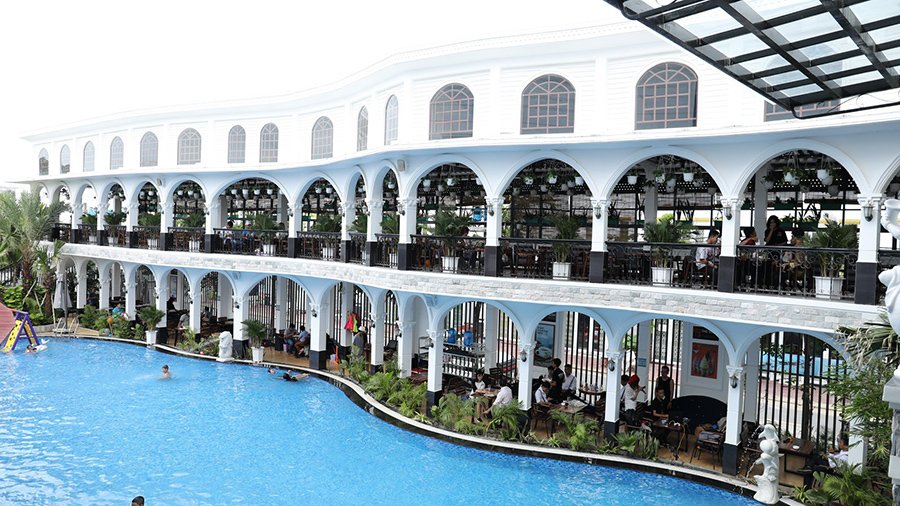 T&ograve;a nh&agrave; lớn hai tầng m&agrave;u trắng với kiến tr&uacute;c v&ograve;m, ph&iacute;a trước l&agrave; hồ bơi xanh v&agrave; một số người đang bơi lội.