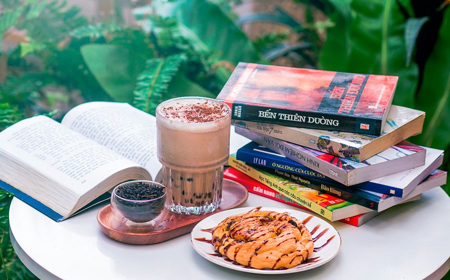 S&aacute;ch, c&agrave; ph&ecirc;, b&aacute;nh ngọt tr&ecirc;n b&agrave;n, xung quanh l&agrave; c&acirc;y xanh.