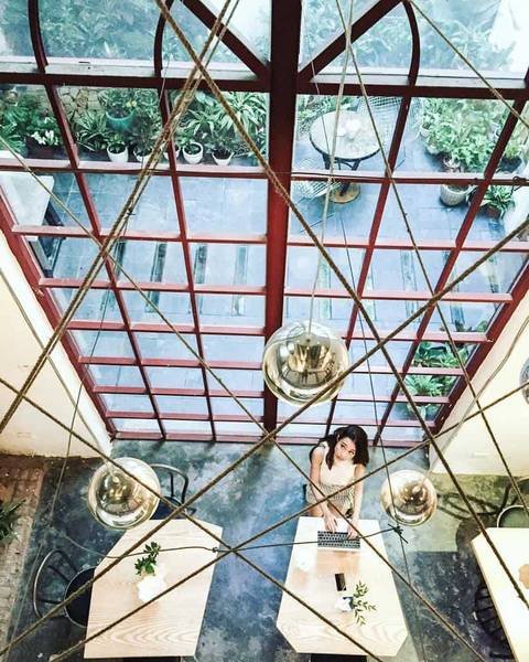 Qu&aacute;n c&agrave; ph&ecirc; nh&igrave;n từ tr&ecirc;n cao với cửa sổ lớn, một người ngồi l&agrave;m việc tr&ecirc;n m&aacute;y t&iacute;nh x&aacute;ch tay, xung quanh c&oacute; c&acirc;y xanh.