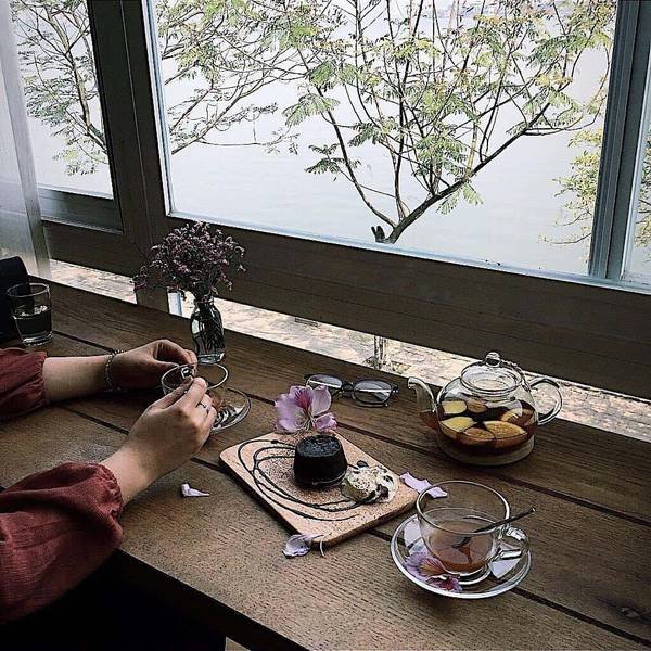 Người ngồi b&ecirc;n b&agrave;n gỗ c&oacute; ấm tr&agrave;, t&aacute;ch tr&agrave; v&agrave; b&aacute;nh ngọt, nh&igrave;n ra cửa sổ với cảnh c&acirc;y v&agrave; s&ocirc;ng.