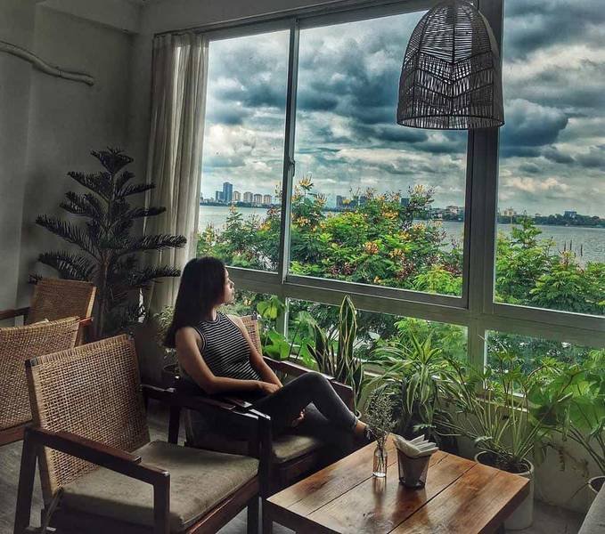 Người ngồi trong qu&aacute;n c&agrave; ph&ecirc;, nh&igrave;n ra ngo&agrave;i cửa sổ lớn với cảnh c&acirc;y cối v&agrave; bầu trời nhiều m&acirc;y.