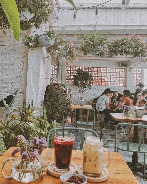Qu&aacute;n c&agrave; ph&ecirc; với kh&ocirc;ng gian ngo&agrave;i trời, b&agrave;n ghế gỗ, c&acirc;y xanh trang tr&iacute;, v&agrave; đồ uống tr&ecirc;n b&agrave;n trước mặt.
