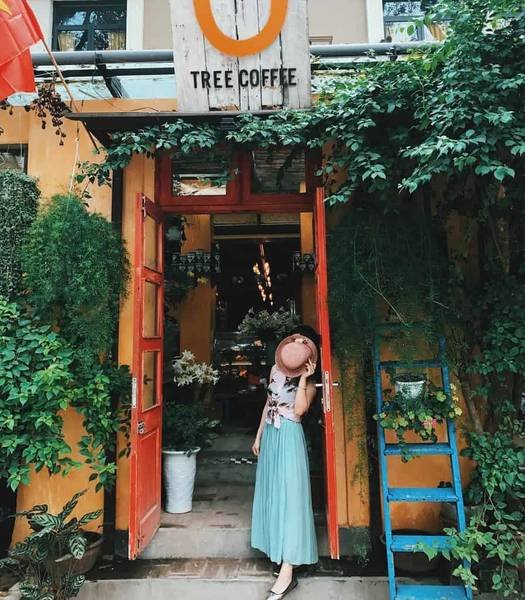 Người đứng trước qu&aacute;n c&agrave; ph&ecirc; với cửa m&agrave;u đỏ, xung quanh l&agrave; c&acirc;y xanh, cầm n&oacute;n che mặt, mặc v&aacute;y d&agrave;i.