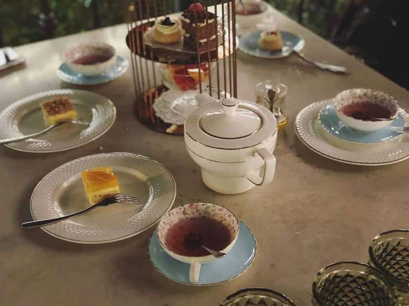 B&agrave;n tiệc tr&agrave; với t&aacute;ch tr&agrave;, b&aacute;nh ngọt tr&ecirc;n đĩa v&agrave; khay nhiều tầng. B&igrave;nh tr&agrave; trắng ở giữa.