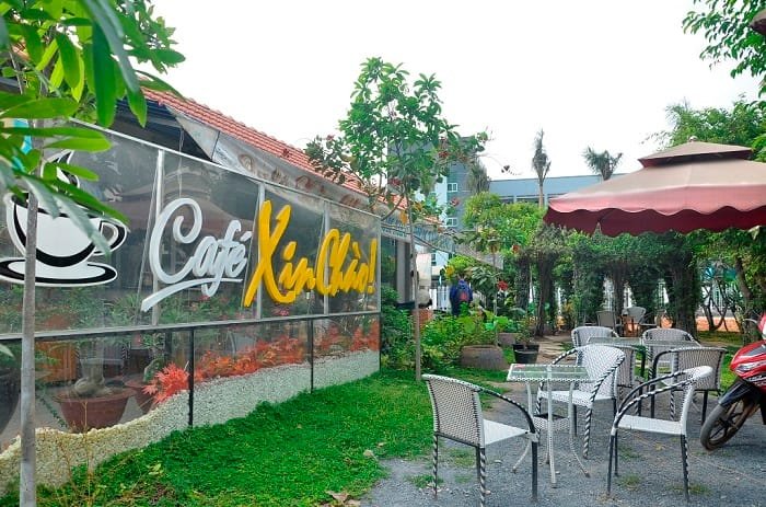 Quán cà phê ngoài trời với bàn ghế, cây xanh và ô che nắng, có biển hiệu "Café Xin Chào" bên cạnh.