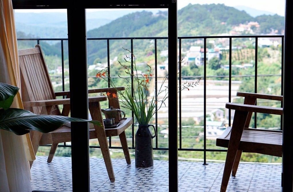 Hai ghế gỗ tr&ecirc;n ban c&ocirc;ng với b&igrave;nh hoa, nh&igrave;n ra cảnh n&uacute;i non xanh tươi qua khung cửa k&iacute;nh.