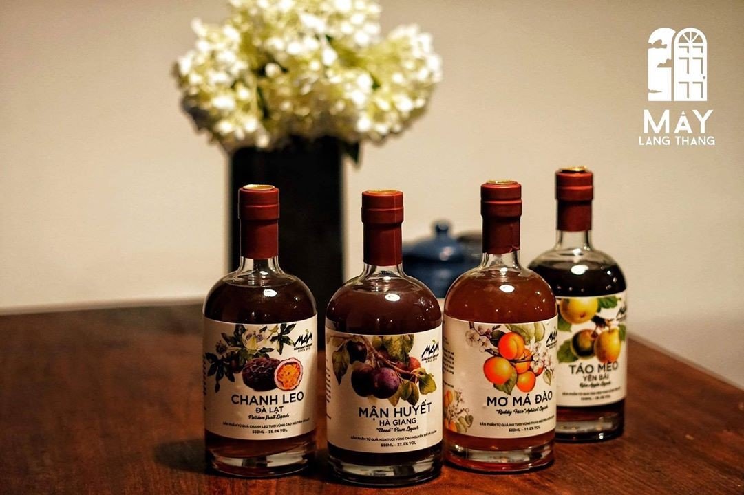 Bốn chai rượu tr&aacute;i c&acirc;y tr&ecirc;n b&agrave;n gỗ, ph&iacute;a sau l&agrave; b&igrave;nh hoa trắng.