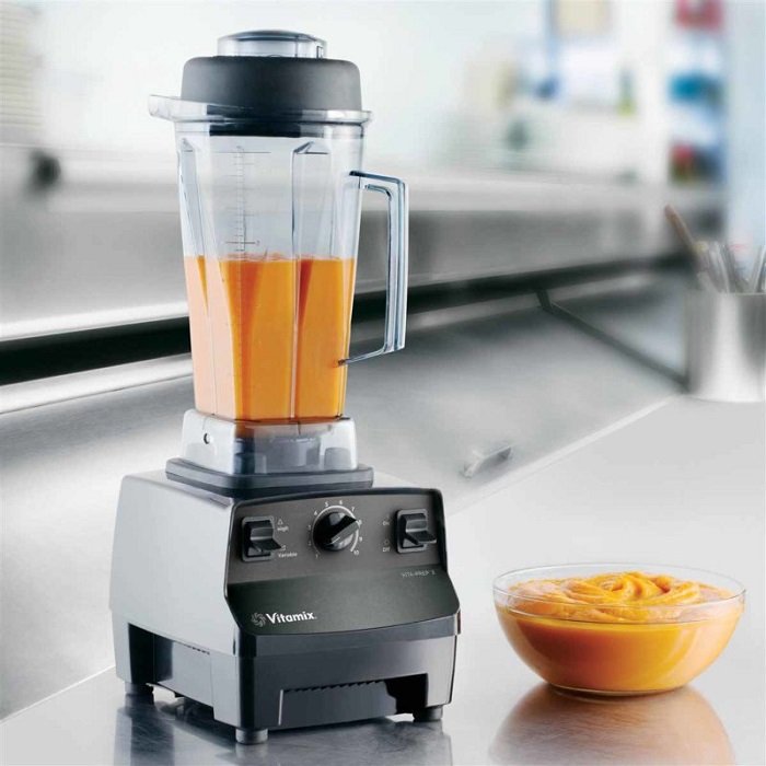 Máy xay sinh tố chứa nước ép màu cam, đặt trên bề mặt bếp, cạnh đó là một bát đựng hỗn hợp tương tự.