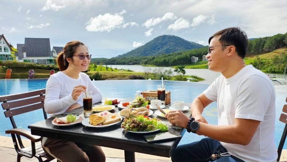 Hai người ngồi ăn s&aacute;ng b&ecirc;n hồ bơi v&ocirc; cực với cảnh n&uacute;i non v&agrave; nh&agrave; cửa xa xa, trời quang đ&atilde;ng.