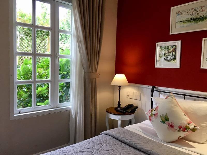 Ph&ograve;ng ngủ với tường đỏ, giường c&oacute; gối hoa, đ&egrave;n b&agrave;n, cửa sổ nh&igrave;n ra c&acirc;y xanh, v&agrave; tranh treo tường.