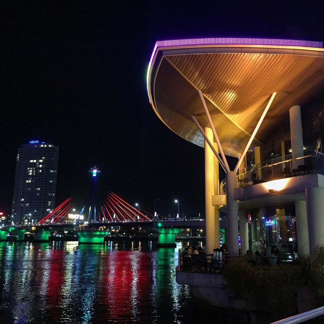 memory-lounge-da-nang-chiec-la-khong-lo-ben-canh-song-han-7-1636482663 Cảnh đêm với cây cầu và tòa nhà hiện đại, ánh sáng màu sắc phản chiếu trên mặt nước yên tĩnh.