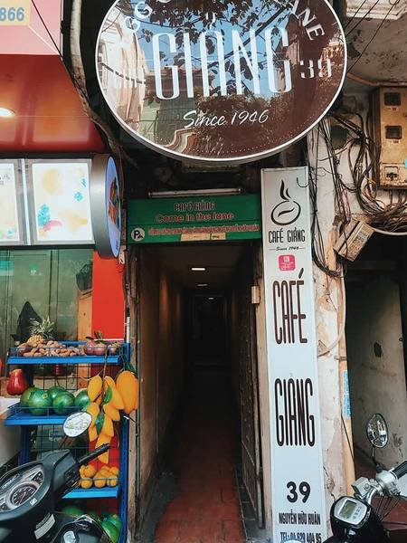 Lối v&agrave;o qu&aacute;n c&agrave; ph&ecirc; nhỏ, b&ecirc;n cạnh c&oacute; bảng hiệu v&agrave; xe m&aacute;y đậu, xung quanh c&oacute; cửa h&agrave;ng tr&aacute;i c&acirc;y.