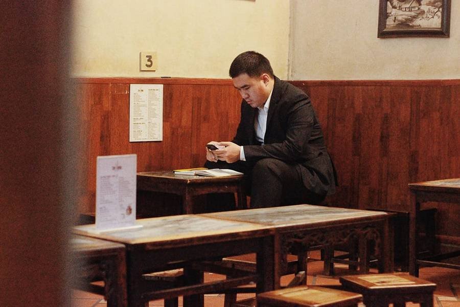 Người đ&agrave;n &ocirc;ng ngồi trong qu&aacute;n c&agrave; ph&ecirc;, mặc &acirc;u phục, tập trung nh&igrave;n v&agrave;o điện thoại, xung quanh l&agrave; b&agrave;n ghế gỗ.