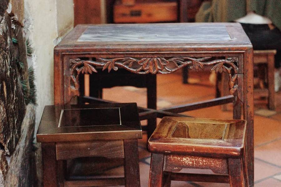 B&agrave;n gỗ chạm khắc hoa văn tinh xảo, k&egrave;m hai ghế gỗ nhỏ, đặt tr&ecirc;n s&agrave;n gạch m&agrave;u n&acirc;u trong kh&ocirc;ng gian cổ k&iacute;nh.