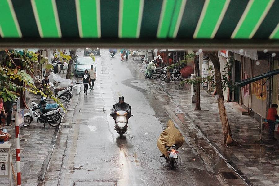 Con đường nhỏ ướt mưa, người d&acirc;n đi xe m&aacute;y v&agrave; đi bộ, c&acirc;y xanh b&ecirc;n vỉa h&egrave;, m&aacute;i hi&ecirc;n sọc xanh ph&iacute;a tr&ecirc;n.
