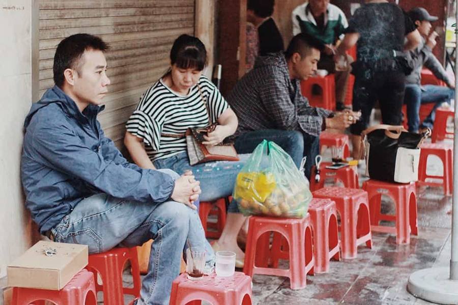 Người ngồi tr&ecirc;n ghế nhựa nhỏ cạnh vỉa h&egrave;, trước cửa h&agrave;ng đ&oacute;ng k&iacute;n, với một t&uacute;i nhựa đựng đồ b&ecirc;n cạnh.