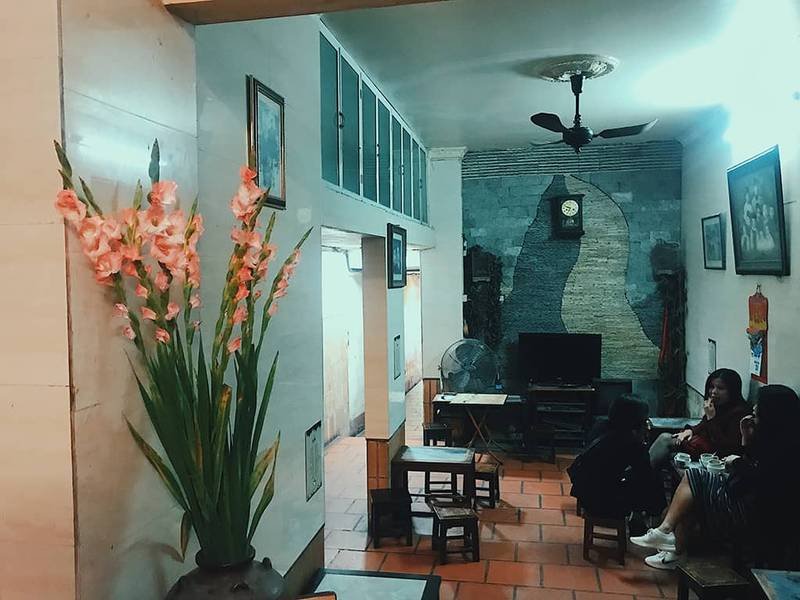 Ph&ograve;ng kh&aacute;ch với b&agrave;n ghế gỗ, b&igrave;nh hoa, quạt trần v&agrave; v&agrave;i người tr&ograve; chuyện, tường trang tr&iacute; tranh v&agrave; đồng hồ.