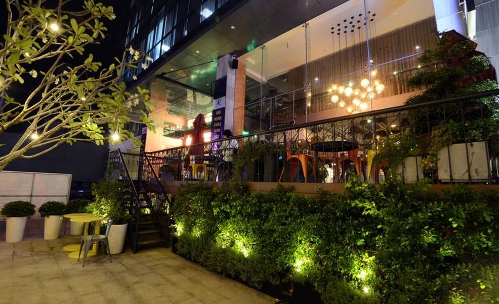 Qu&aacute;n c&agrave; ph&ecirc; với cửa k&iacute;nh, b&agrave;n ghế ngo&agrave;i trời, c&acirc;y cối v&agrave; &aacute;nh s&aacute;ng lung linh v&agrave;o ban đ&ecirc;m.