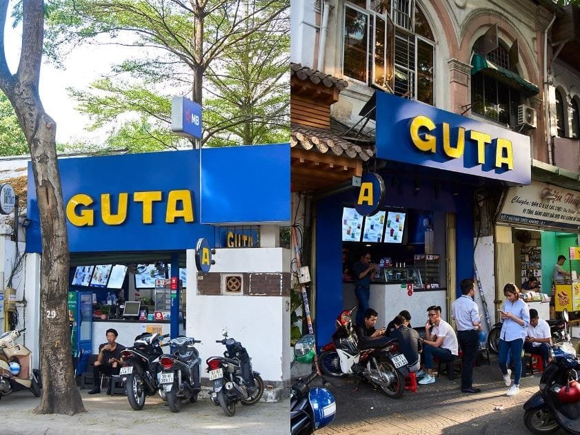 Top 32 thương hiệu nhượng quyền cafe siêu lợi nhuận 23 Hai cửa hàng với biển hiệu "GUTA", có xe máy đỗ phía trước và cây xanh bên cạnh.