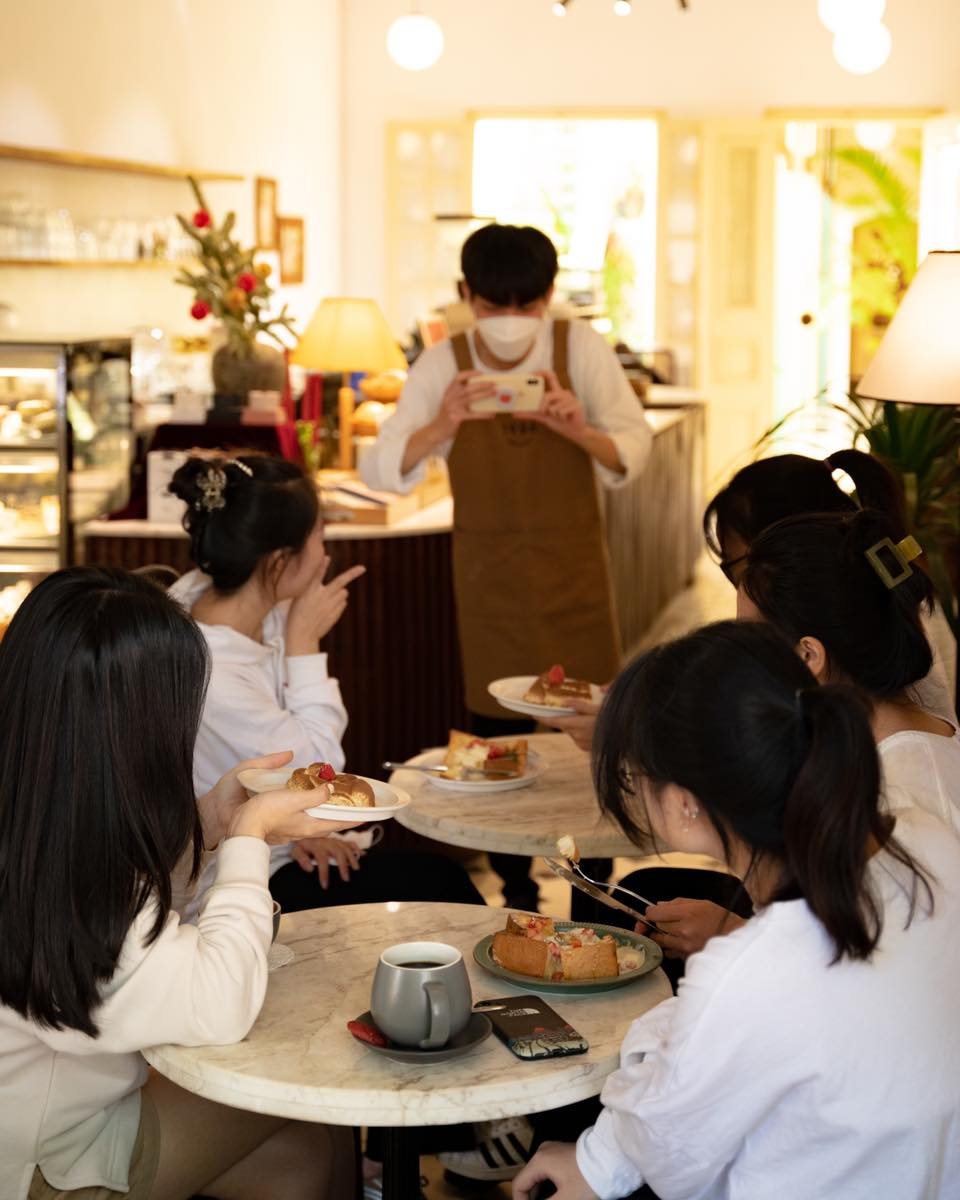 Nh&oacute;m người ngồi trong qu&aacute;n c&agrave; ph&ecirc;, thưởng thức b&aacute;nh ngọt. Nh&acirc;n vi&ecirc;n đang chụp ảnh bằng điện thoại.