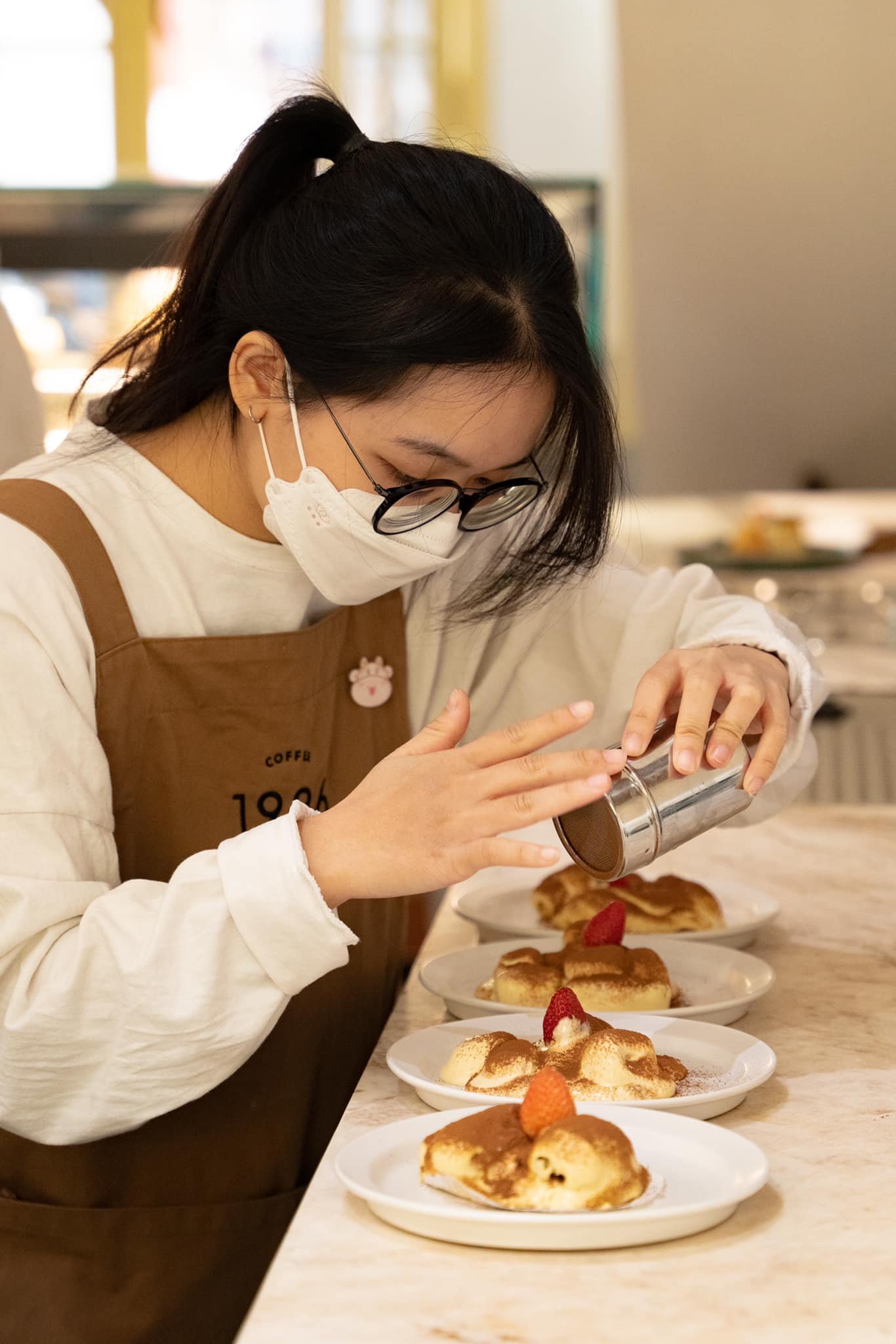 Người l&agrave;m b&aacute;nh đang trang tr&iacute; m&oacute;n tr&aacute;ng miệng với bột cacao, tr&ecirc;n b&agrave;n c&oacute; nhiều đĩa b&aacute;nh xếp h&agrave;ng.