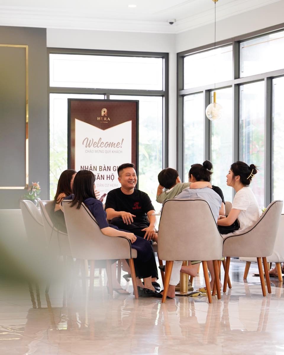 Một nh&oacute;m người ngồi quanh b&agrave;n tr&ograve; chuyện trong một căn ph&ograve;ng s&aacute;ng, c&oacute; bảng ch&agrave;o mừng ph&iacute;a sau.