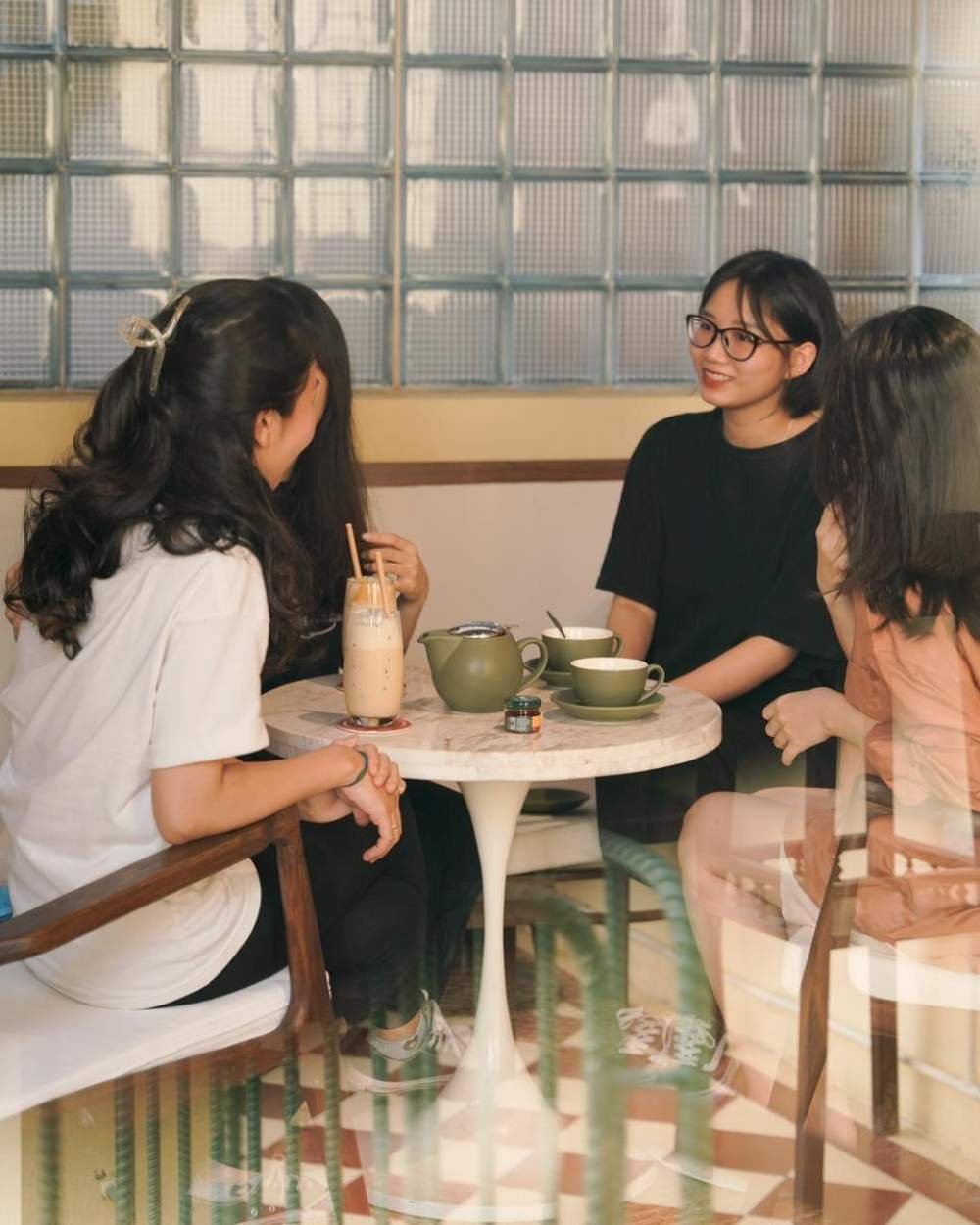 Bốn người ngồi quanh b&agrave;n tr&ograve;n trong qu&aacute;n c&agrave; ph&ecirc;, tr&ograve; chuyện v&agrave; uống nước. Tr&ecirc;n b&agrave;n c&oacute; tr&agrave; v&agrave; cốc nước.