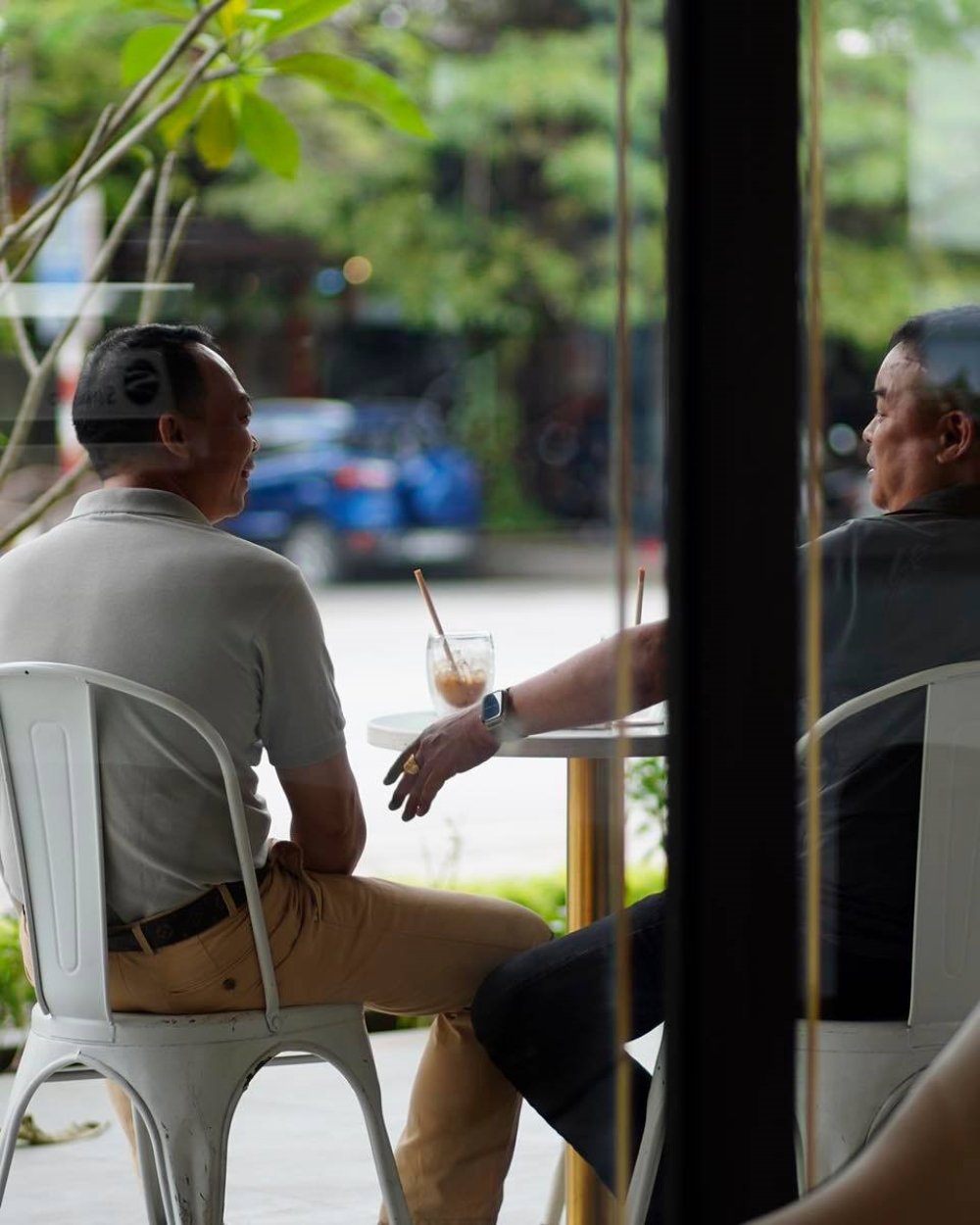 Hai người ngồi tr&ograve; chuyện tại qu&aacute;n c&agrave; ph&ecirc;, b&ecirc;n ngo&agrave;i c&oacute; c&acirc;y xanh v&agrave; xe &ocirc; t&ocirc; đậu.