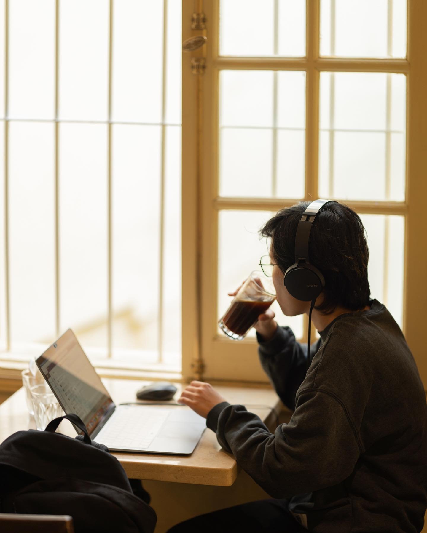 Người ngồi cạnh cửa sổ, đeo tai nghe, sử dụng laptop v&agrave; uống c&agrave; ph&ecirc; trong ly thủy tinh.