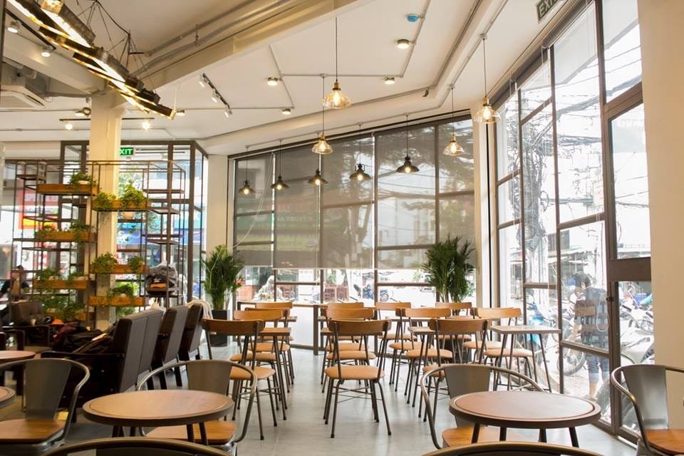 Top 32 thương hiệu nhượng quyền cafe siêu lợi nhuận 26 Quán cà phê sáng sủa với bàn ghế gỗ, cửa kính lớn, và cây xanh trang trí, tạo không gian thoáng đãng.