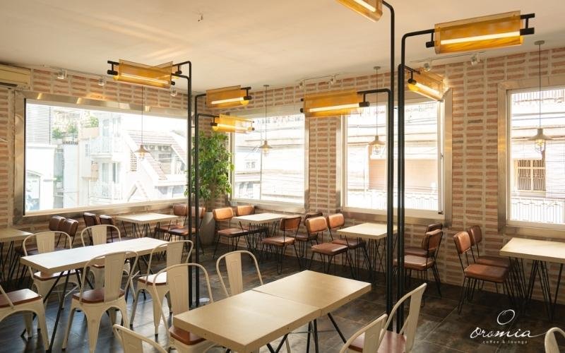 Quán cà phê với bàn ghế gỗ, đèn trang trí và cửa sổ lớn nhìn ra khung cảnh bên ngoài, tạo không gian sáng sủa.