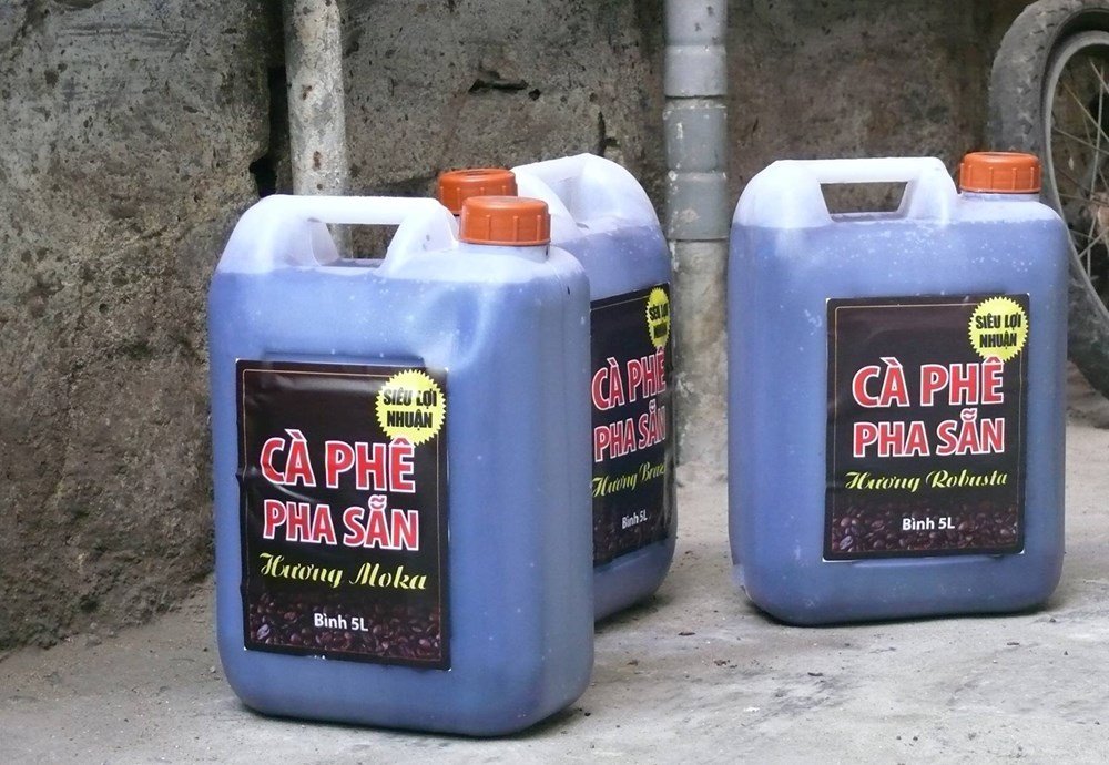 Ba bình nhựa lớn chứa chất lỏng có nhãn "Cà Phê Pha Sẵn", đặt trên nền xi măng gần một bức tường.