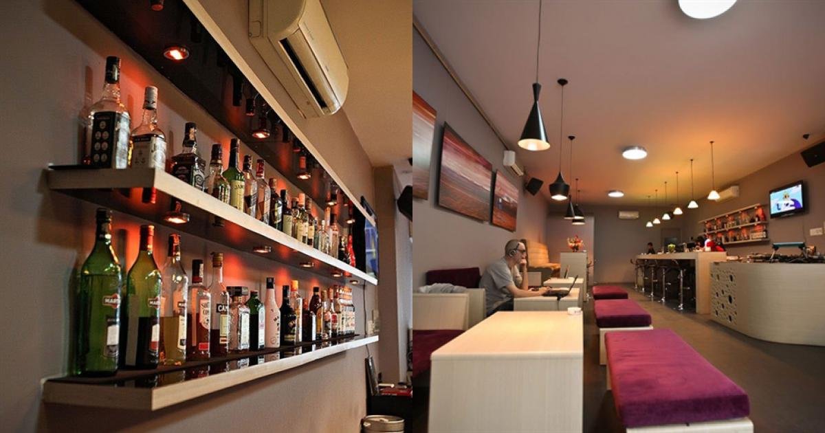 Quầy bar hiện đại với kệ rượu, bàn ghế gỗ, ánh sáng ấm áp và một người sử dụng máy tính xách tay.