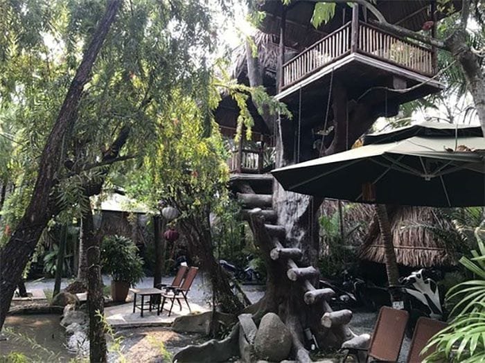 Gợi ý 14 quán cà phê chòi mát mẻ, thư giãn tại TPHCM 5 Ngôi nhà trên cây với cầu thang gỗ và khu vườn xanh mát, có bàn ghế và ô che nắng.