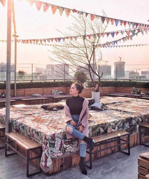 Người ngồi tr&ecirc;n s&acirc;n thượng trang tr&iacute; với cờ m&agrave;u sắc v&agrave; c&acirc;y cảnh, ph&iacute;a sau l&agrave; cảnh th&agrave;nh phố l&uacute;c ho&agrave;ng h&ocirc;n.