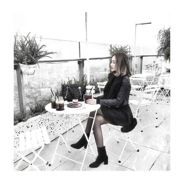 Người ngồi tại qu&aacute;n c&agrave; ph&ecirc; ngo&agrave;i trời, b&agrave;n trắng, xung quanh c&oacute; c&acirc;y xanh, với đồ uống v&agrave; t&uacute;i x&aacute;ch tr&ecirc;n b&agrave;n.