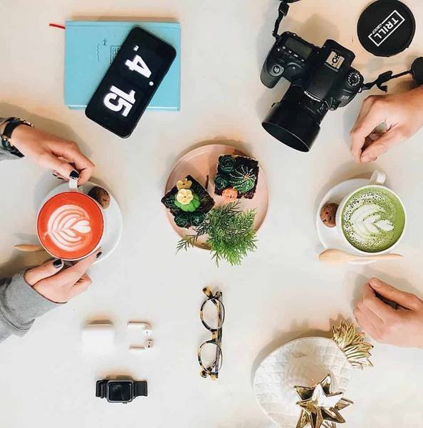 Hai người cầm t&aacute;ch đồ uống tr&ecirc;n b&agrave;n với m&aacute;y ảnh, điện thoại, đồng hồ, v&agrave; đĩa b&aacute;nh trang tr&iacute;.