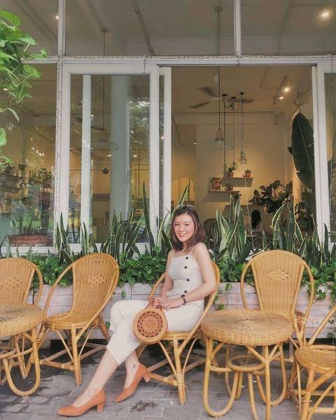 Người phụ nữ ngồi ngo&agrave;i qu&aacute;n c&agrave; ph&ecirc; với ghế m&acirc;y v&agrave; c&acirc;y xanh xung quanh, cầm t&uacute;i tr&ograve;n nhỏ.