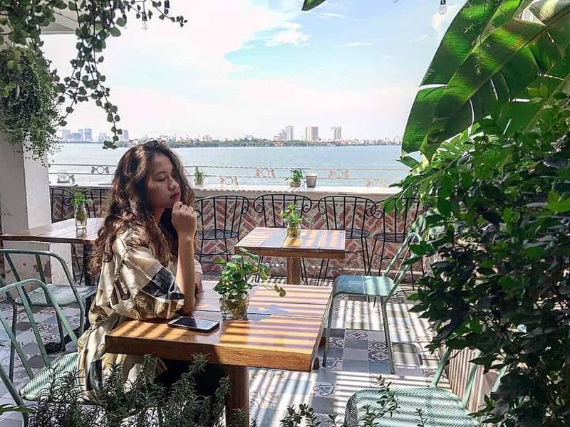 Người ngồi tại qu&aacute;n c&agrave; ph&ecirc; ven hồ, xung quanh c&oacute; nhiều c&acirc;y xanh v&agrave; b&agrave;n ghế, khung cảnh tho&aacute;ng đ&atilde;ng.