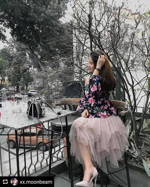 Người phụ nữ ngồi ngo&agrave;i trời b&ecirc;n b&agrave;n c&agrave; ph&ecirc;, mặc v&aacute;y hoa v&agrave; ch&acirc;n v&aacute;y xếp tầng, xung quanh c&oacute; c&acirc;y cối v&agrave; xe cộ.